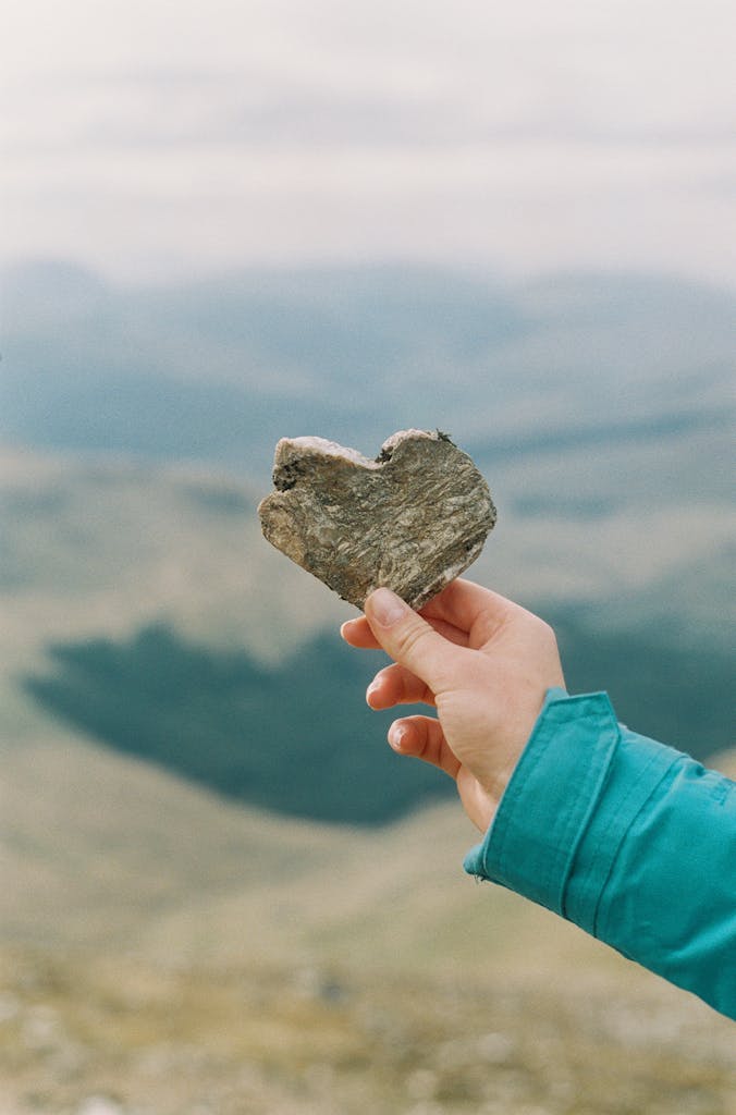 A hand holds a heart-shaped rock against a scenic Scottish Highlands backdrop.